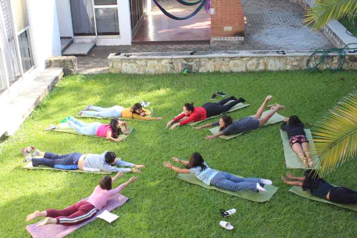 Grupo de nueve mujeres jóvenes acostadas boca abajo, brazos extendidos hacia el frente, en tapete de yoga en jardín.
