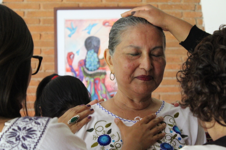 Una mujer de aproximadamente 60 años, tiene los ojos cerrados y una expresión de tranquilidad en el rostro que proviene de las manos de otras mujeres que le tocan a ella sus hombros y su pecho suavemente.